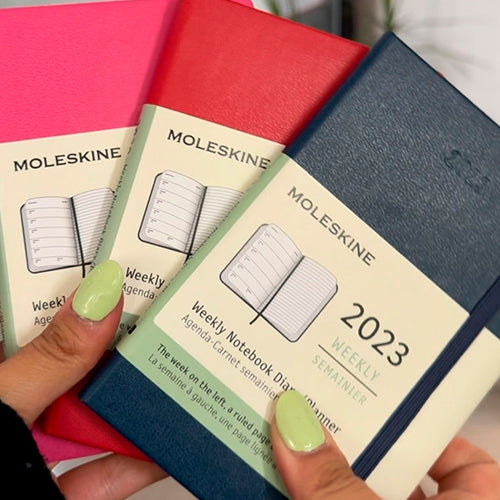 Dos manos sosteniendo tres agendas Moleskine cerradas de colores rosa, rojo y azul, todas con la etiqueta de "Weekly Notebook Diary / Planner" y la fecha "2023".