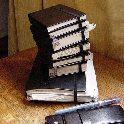 Una pila de cuadernos Moleskine negros de diferentes tamaños, algunos con páginas sobresaliendo y marcadores de cinta, sobre una superficie de madera.